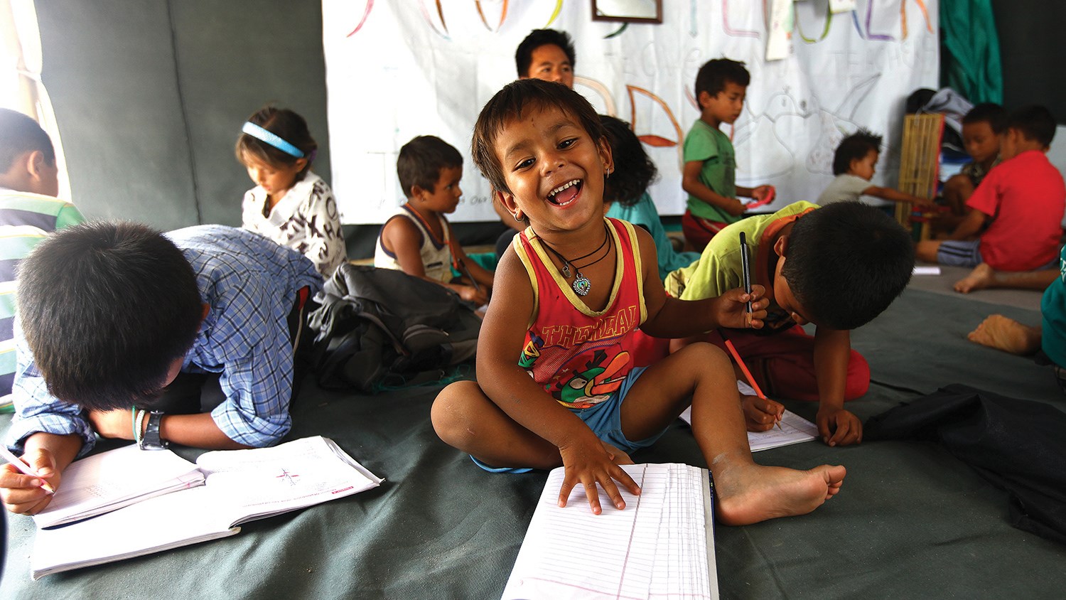 Un jeune enfant est tout sourire en faisant ses devoirs dans un centre pédagogique temporaire financé par l'UNICEF, un espace sûr destiné aux enfants du camp Chuchhepati à Kathmandou, au Népal. Des milliers de gens furent déplacés de leurs foyers par le tremblement de terre d'avril 2015.