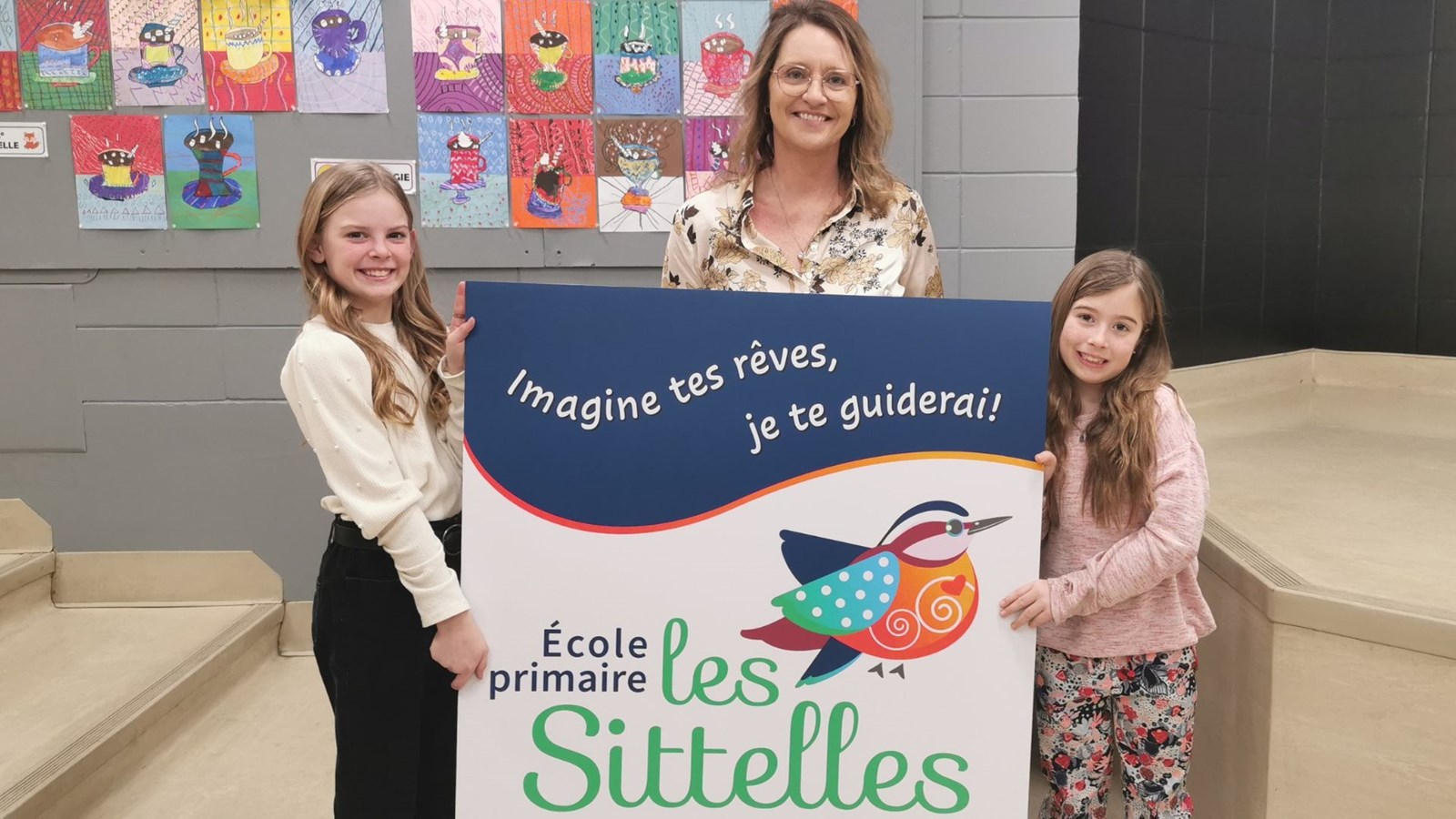 Les élèves Khloe Roy et Eva Lemieux tenant fièrement l'affiche de la nouvelle signature graphique de l'école, réalisée par Chantale Maheux, technicienne en arts graphiques pour le Centre de services scolaires de la Beauce-Etchemin.